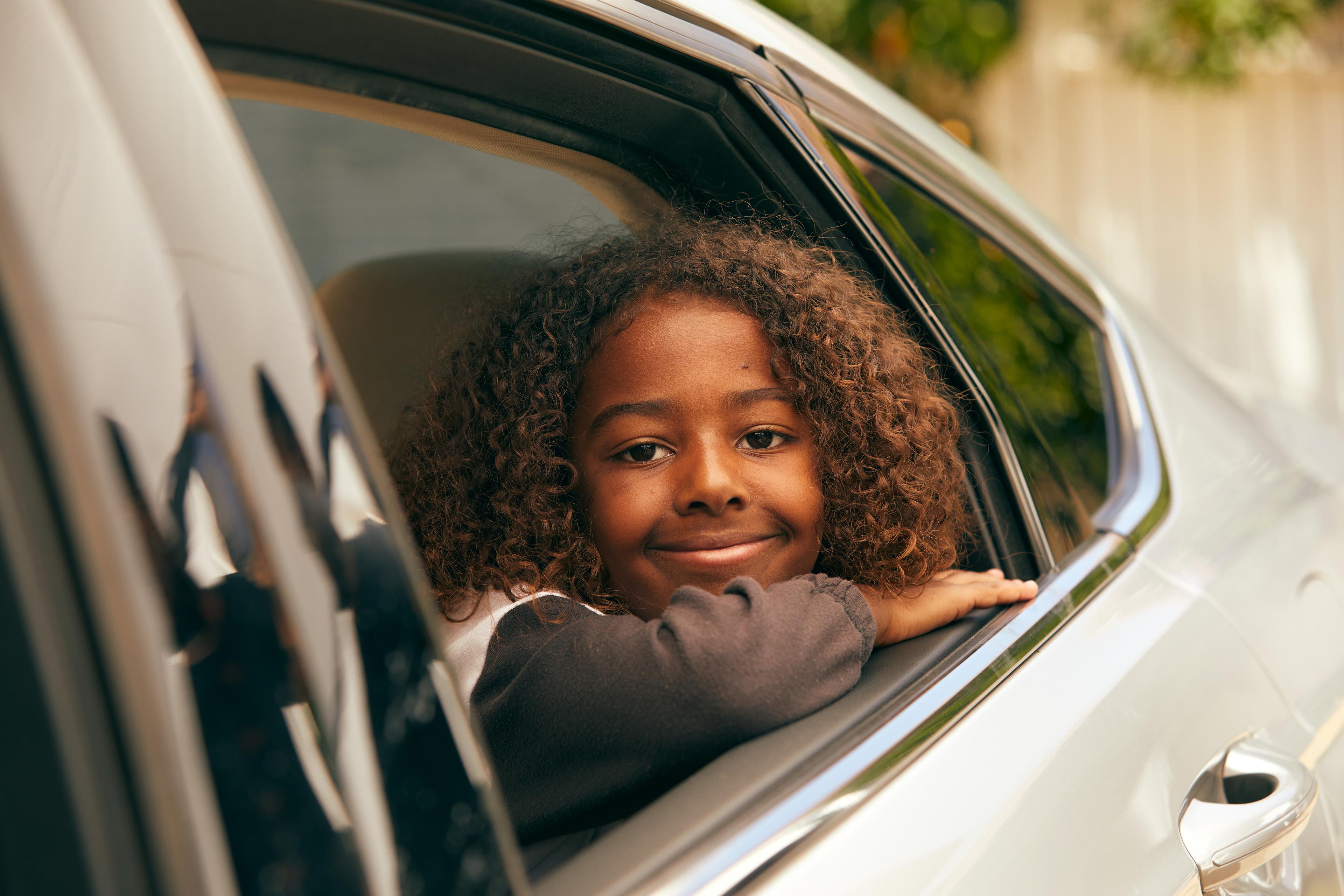 Young passenger with curly hair smiling through car window, wearing dark clothing, natural warm lighting