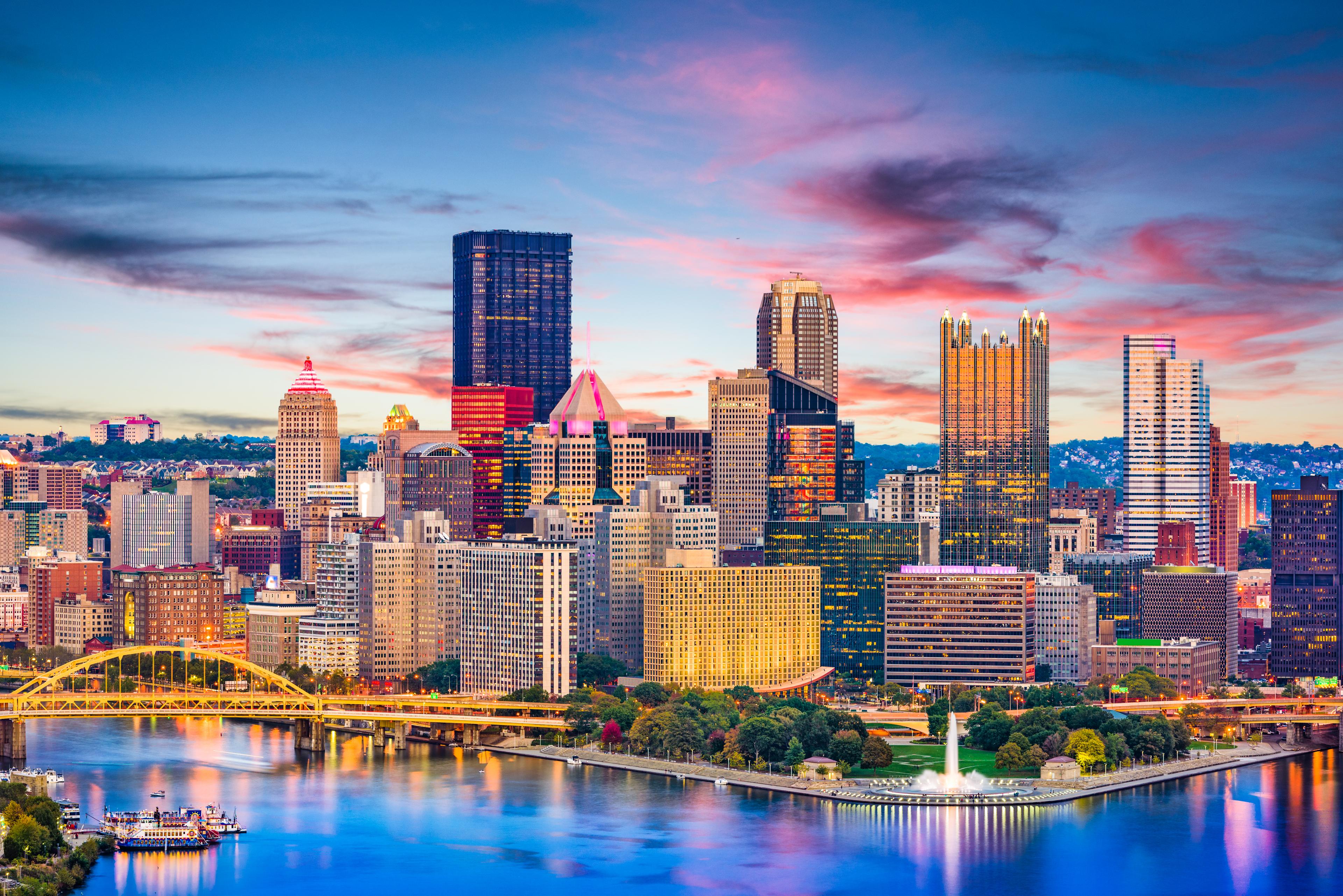 Pittsburgh's downtown skyline reflecting in the Ohio River at sunset, with colorful sky and illuminated skyscrapers
