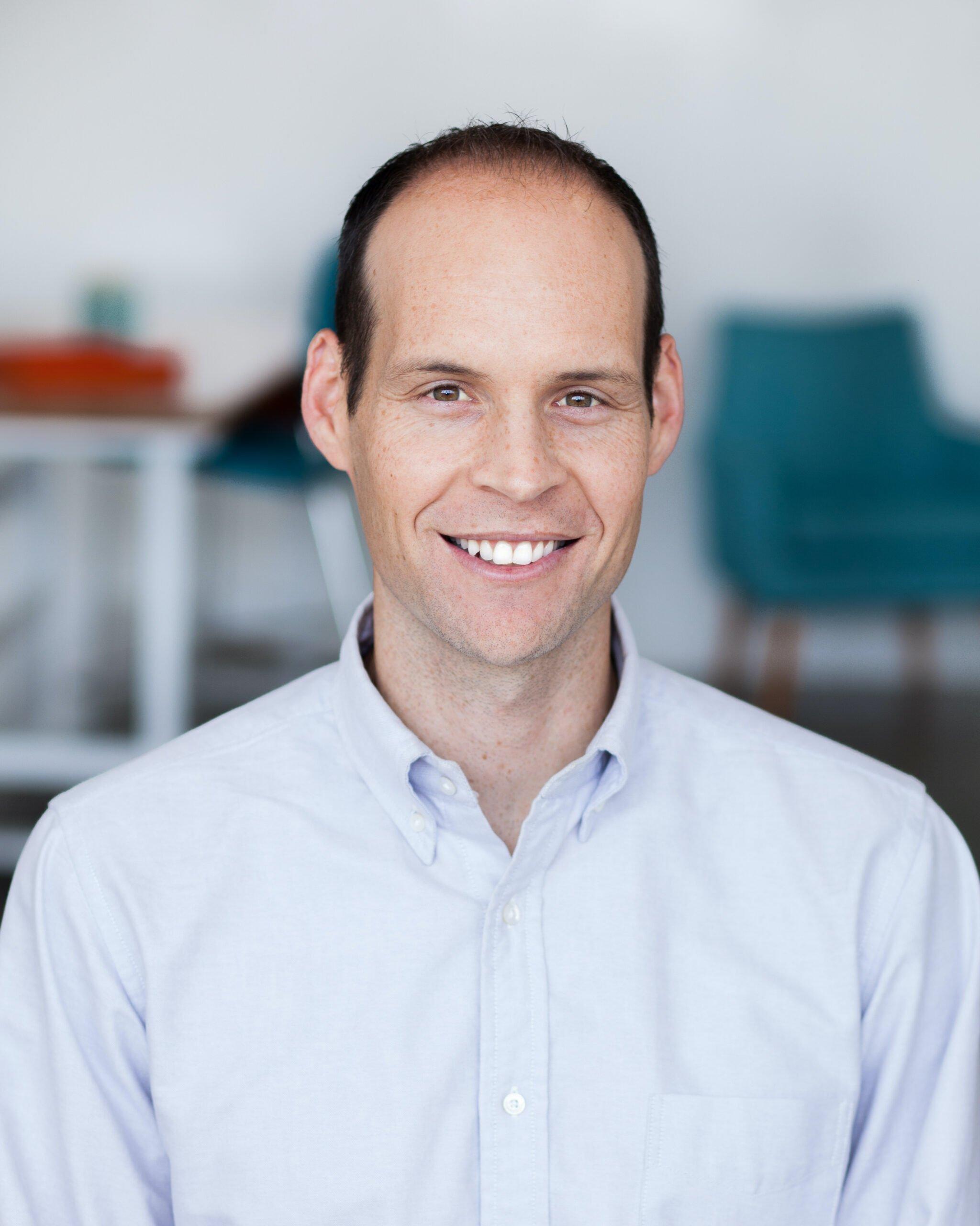 Professional headshot of person in white button-down shirt smiling at camera with office background