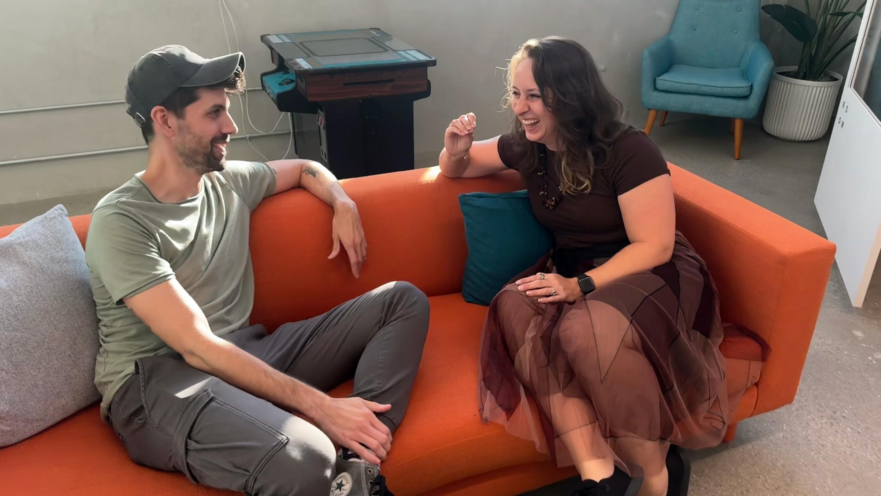 Two people having an animated conversation while sitting on a bright orange couch in a modern office setting with teal accents