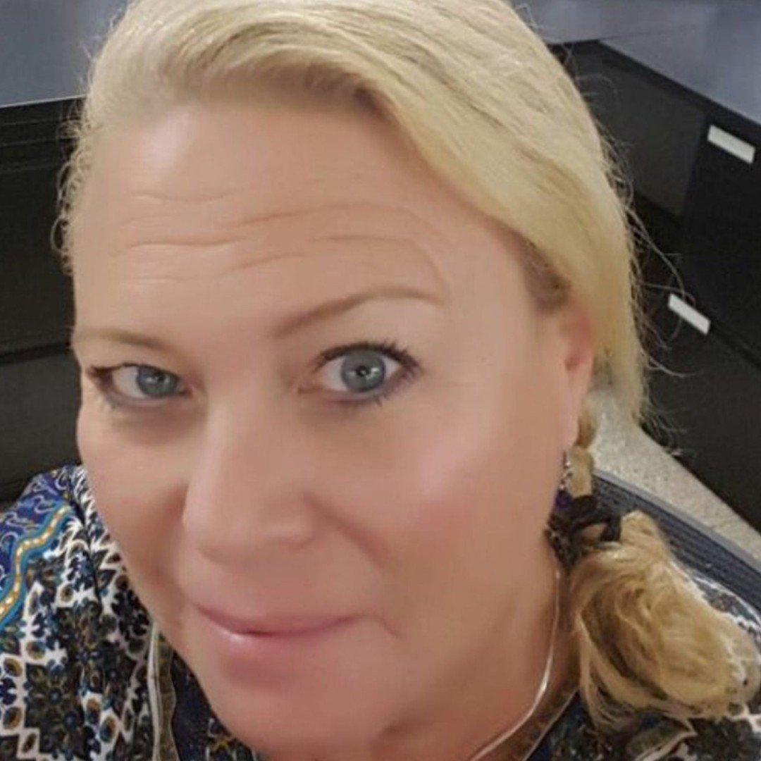 Close-up portrait of a person with blonde hair and blue eyes wearing a patterned blue and white blouse, smiling warmly at camera