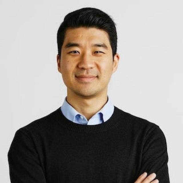 Professional headshot of a person in business casual attire wearing a black sweater over a light blue button-up shirt, with arms crossed and a warm smile