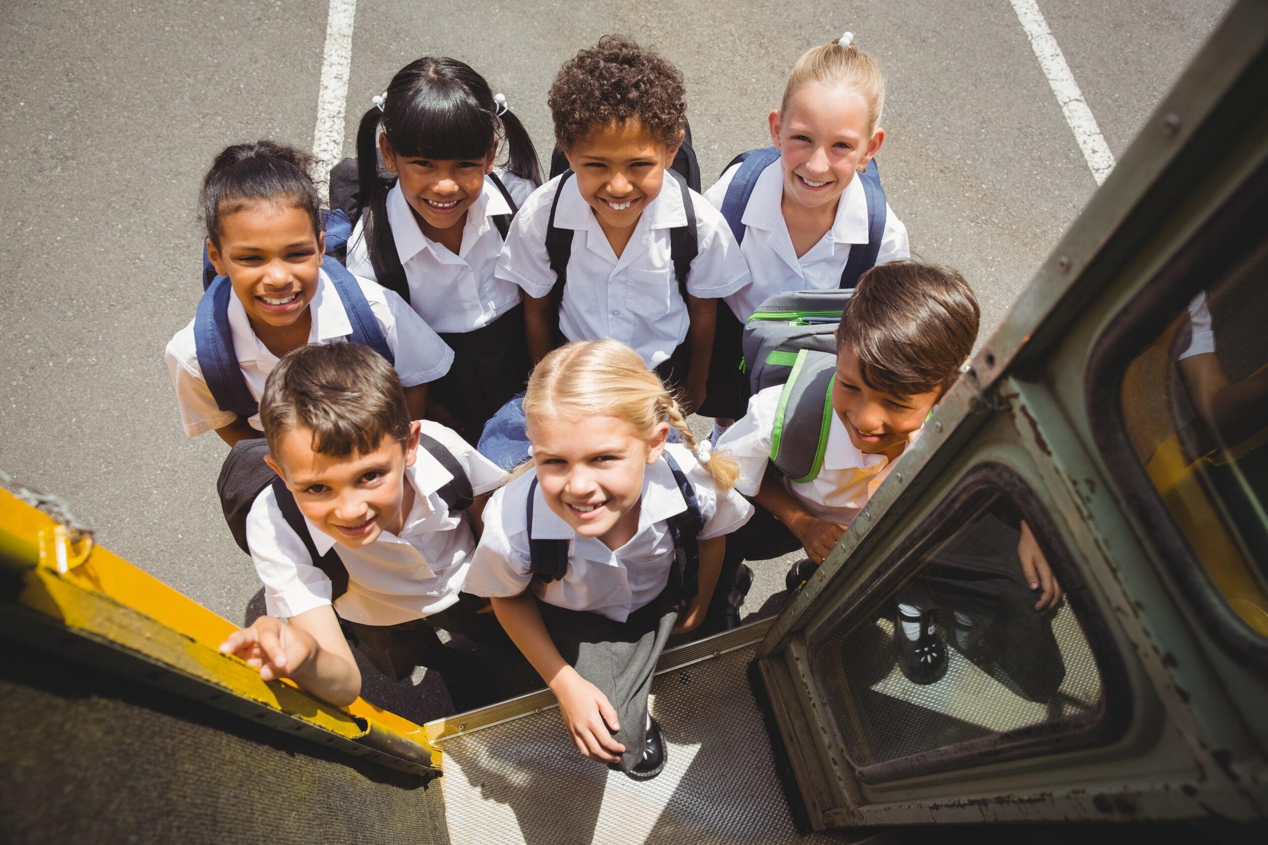 Group of elementary school students in uniforms smiling up at camera while boarding a yellow school bus