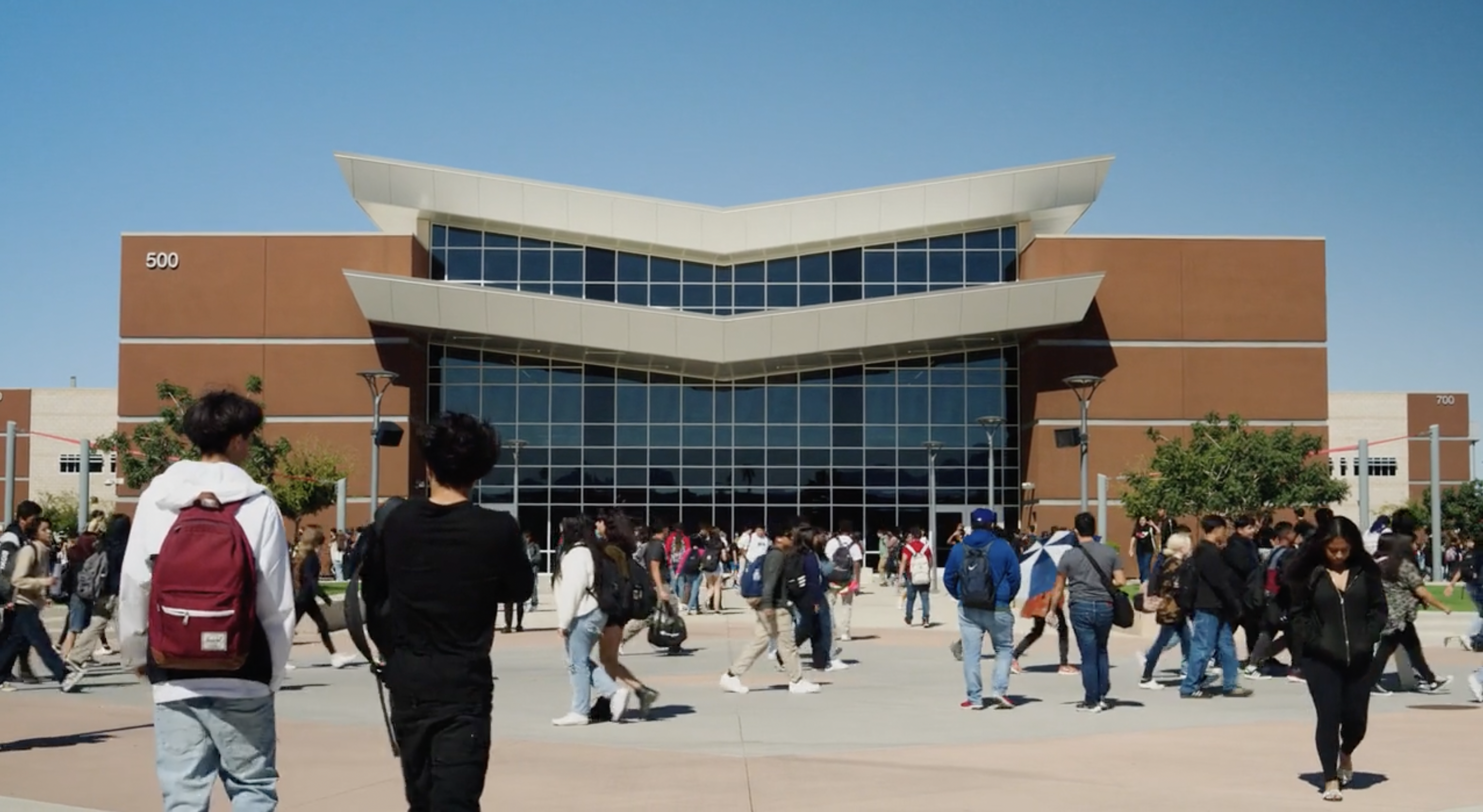 Modern two-story educational building with glass facade and angular roof design, surrounded by students walking between classes