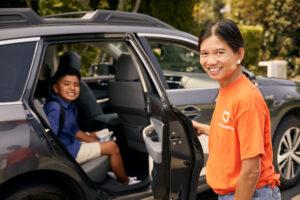 Person in orange t-shirt standing by car door with passenger seated in back seat, both smiling in daytime setting