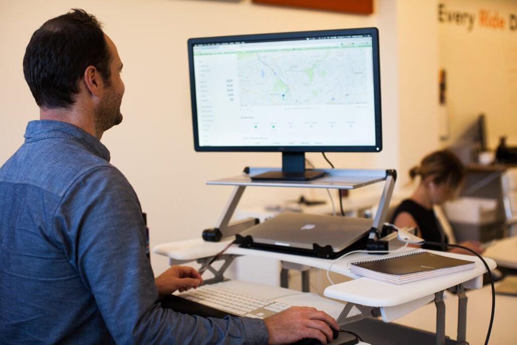 Professional in blue shirt working at adjustable standing desk setup with large monitor displaying map interface