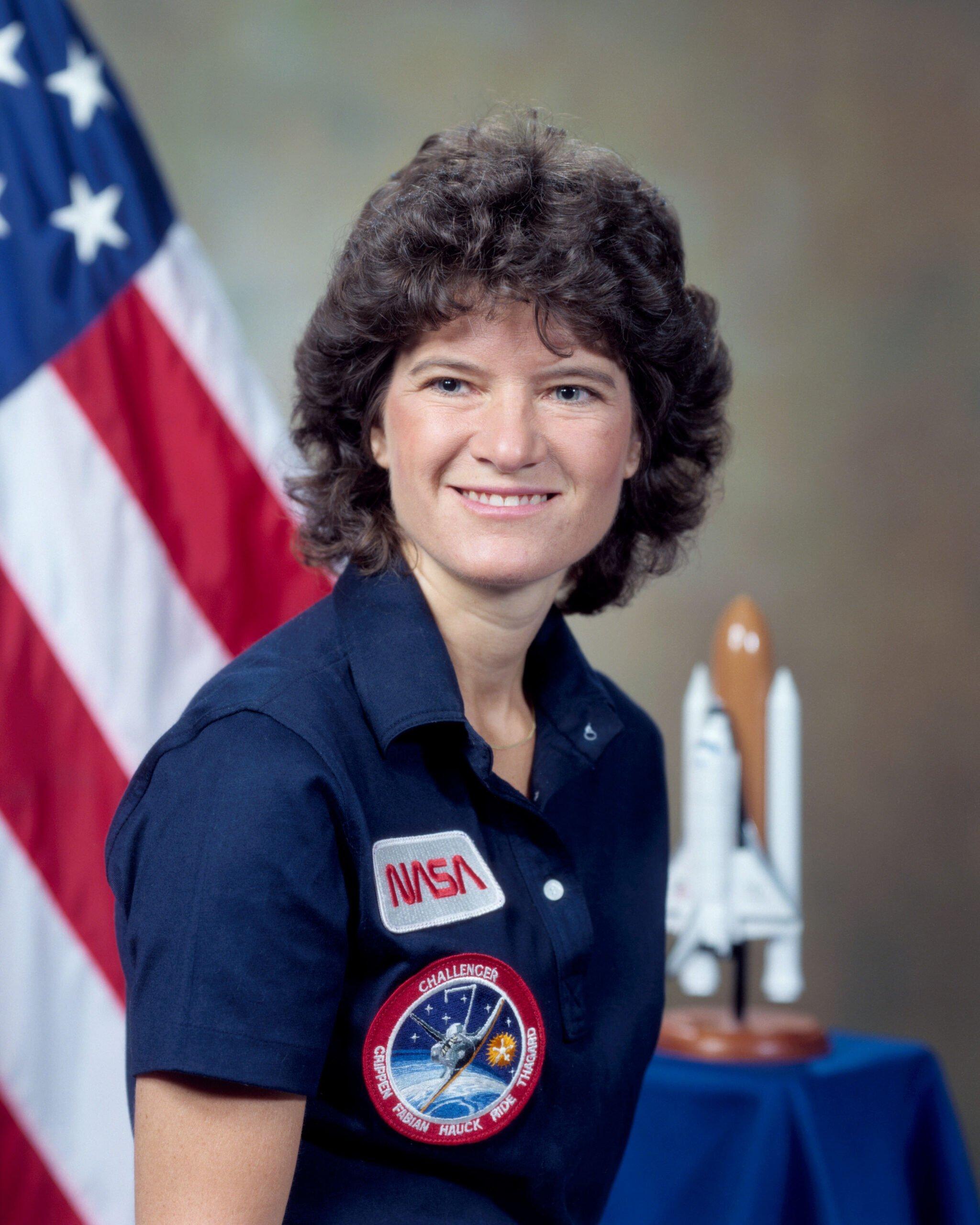 Astronaut in navy NASA uniform with Challenger mission patch posing for official portrait with American flag and shuttle model