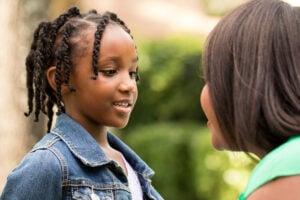 Two people engaged in a friendly conversation outdoors, one wearing a denim jacket with braided hair, the other in a green top