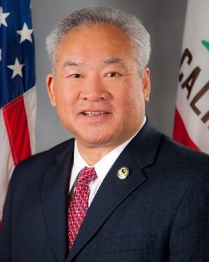 Professional headshot of an official in a dark suit and red tie with US and California state flags in the background
