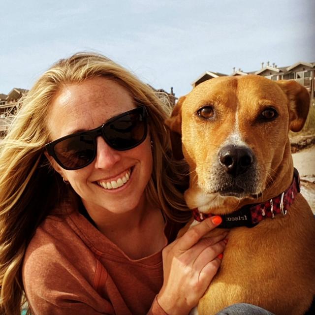 A close-up selfie featuring someone in sunglasses smiling next to a brown mixed breed dog wearing a red collar, with beach houses visible in the background