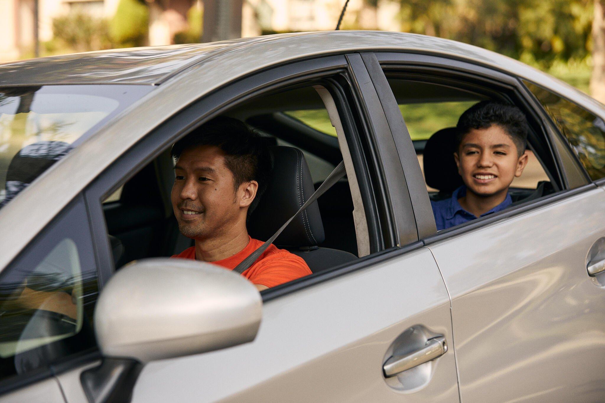 Two people sitting in a silver car, driver in orange shirt and passenger in blue shirt, both smiling and wearing seatbelts