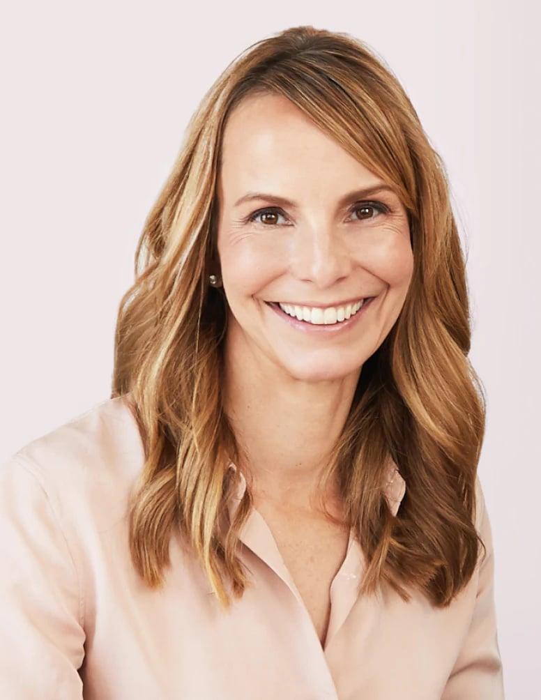 Professional headshot of woman with honey-blonde hair wearing light pink blouse, smiling warmly against soft pink background