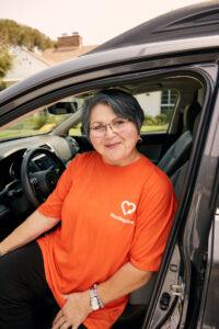 A person wearing an orange company t-shirt sits in the driver's seat of a car, smiling warmly at the camera