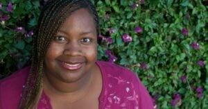 Portrait of a woman in a burgundy top smiling warmly, with green foliage and purple flowers in the background