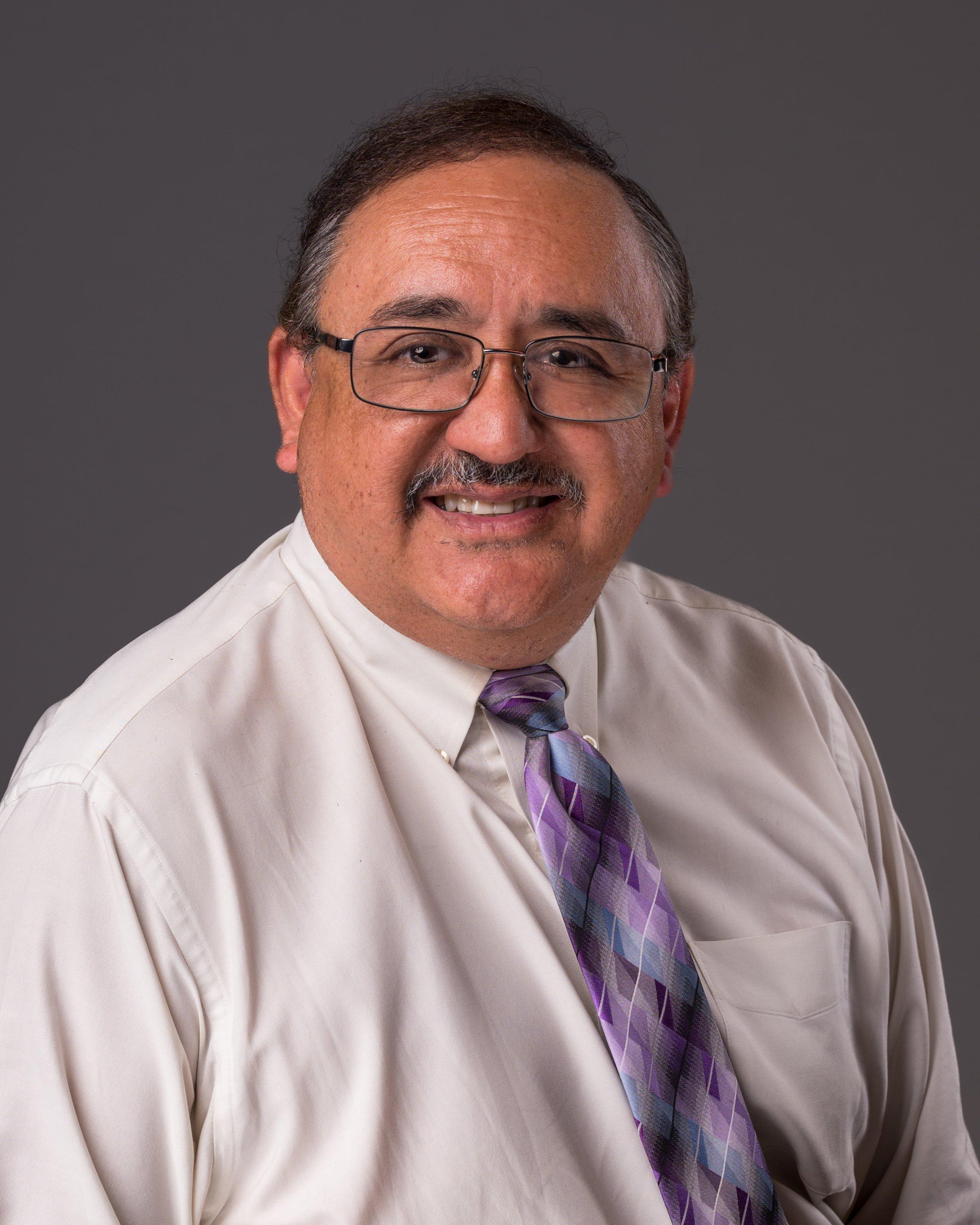 Professional headshot of a middle-aged man wearing glasses, white dress shirt, and purple patterned tie, smiling warmly at camera