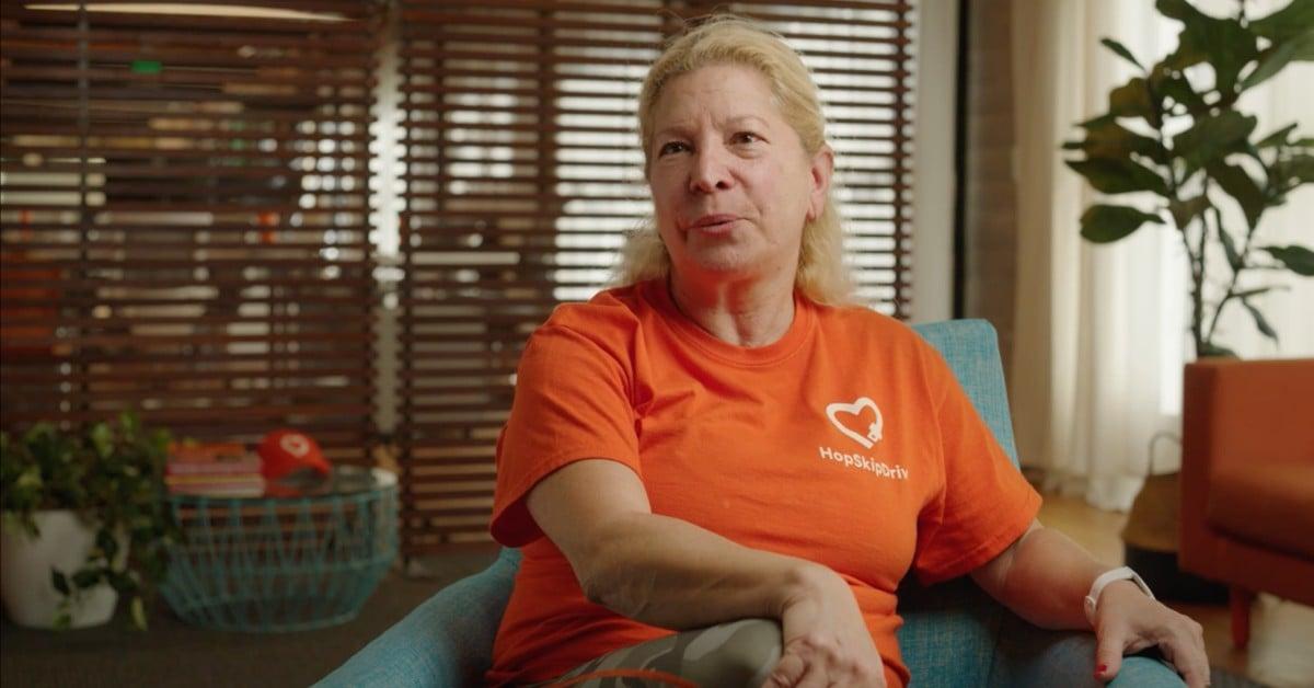 Woman in bright orange HopSkipDrive t-shirt sitting in blue chair during interview, home or office setting with plants in background