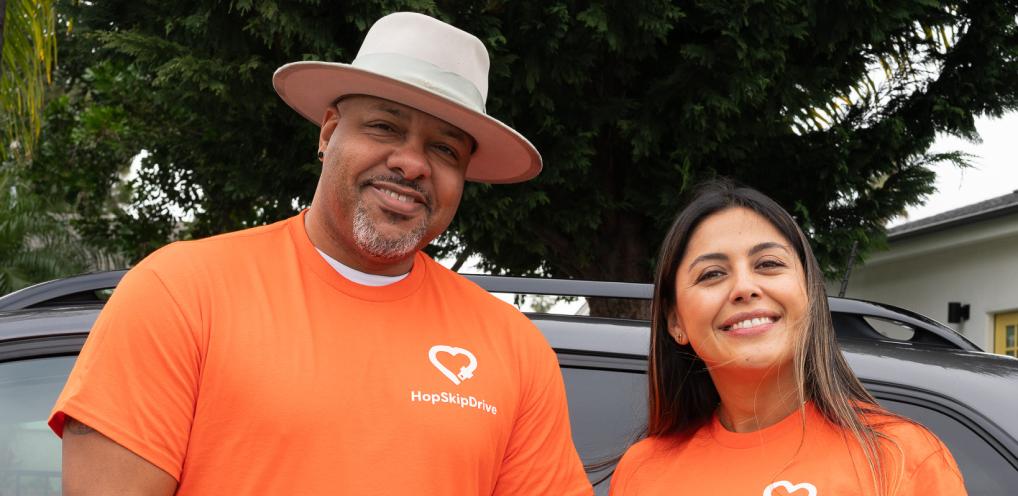Two team members wearing bright orange HopSkipDrive t-shirts with heart logo, one wearing a white hat, standing in front of vehicles