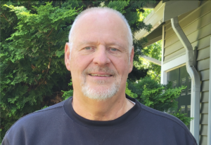 Portrait of a middle-aged man with a beard, wearing a black shirt, standing in front of a house and evergreen shrubs