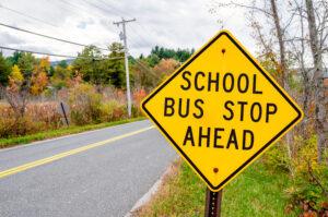 Yellow diamond-shaped school bus stop ahead warning sign along a rural road with autumn foliage in the background