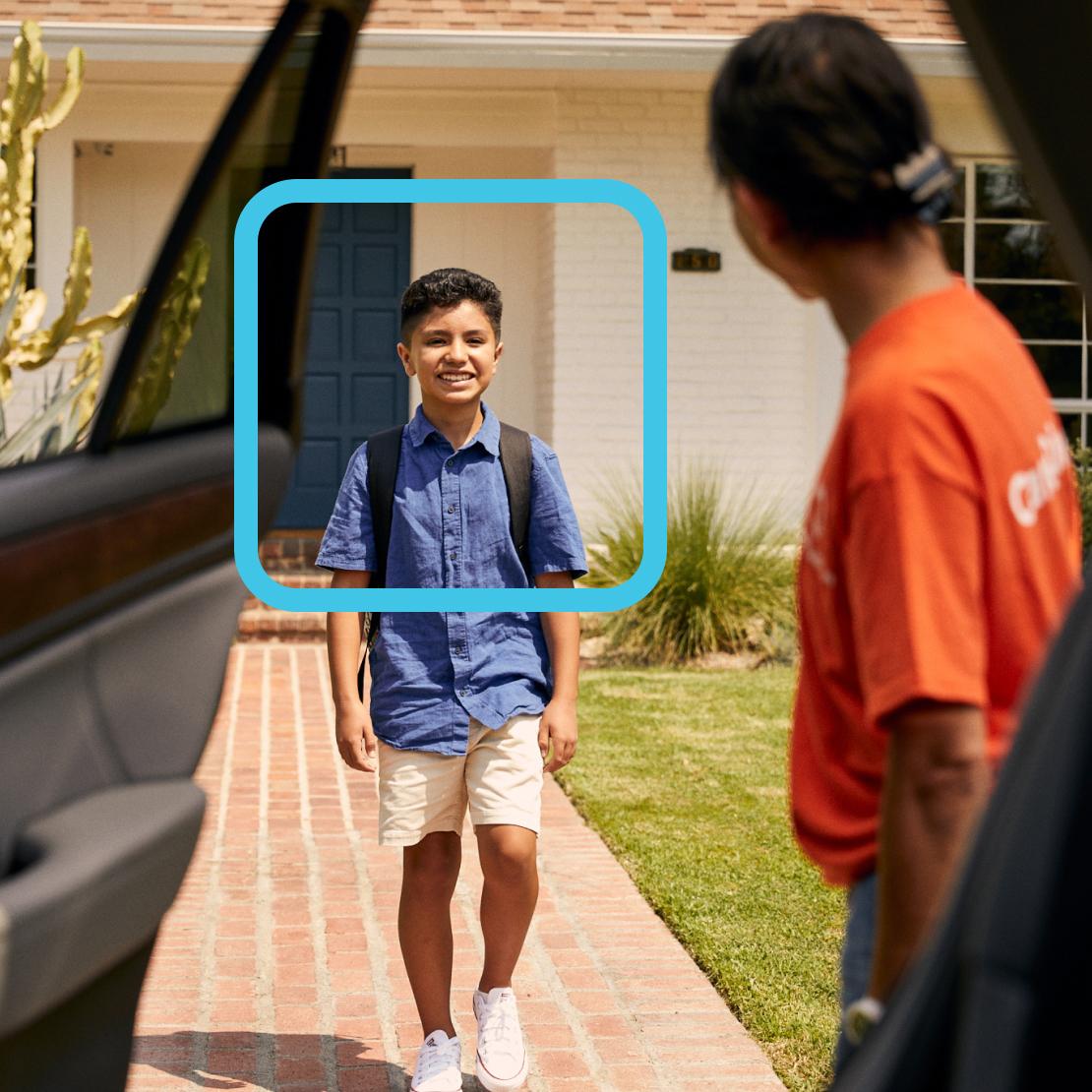 A young student wearing a blue shirt and khaki shorts stands smiling on a brick pathway with a backpack