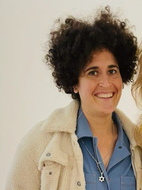Woman with curly black hair smiling warmly, wearing a cream sherpa jacket and blue button-up shirt against a light background