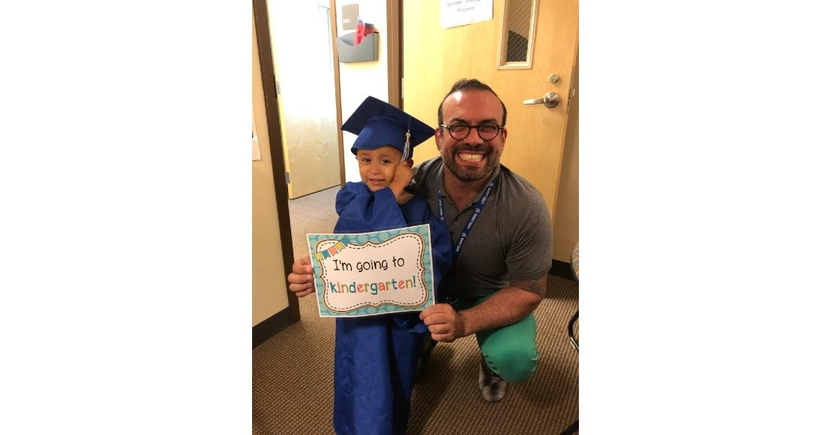 Young student in blue graduation cap and gown holds 'I'm going to kindergarten!' sign while posing with educator
