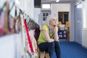 Athletic person in neon yellow top sitting thoughtfully on bench in locker room with sports equipment visible