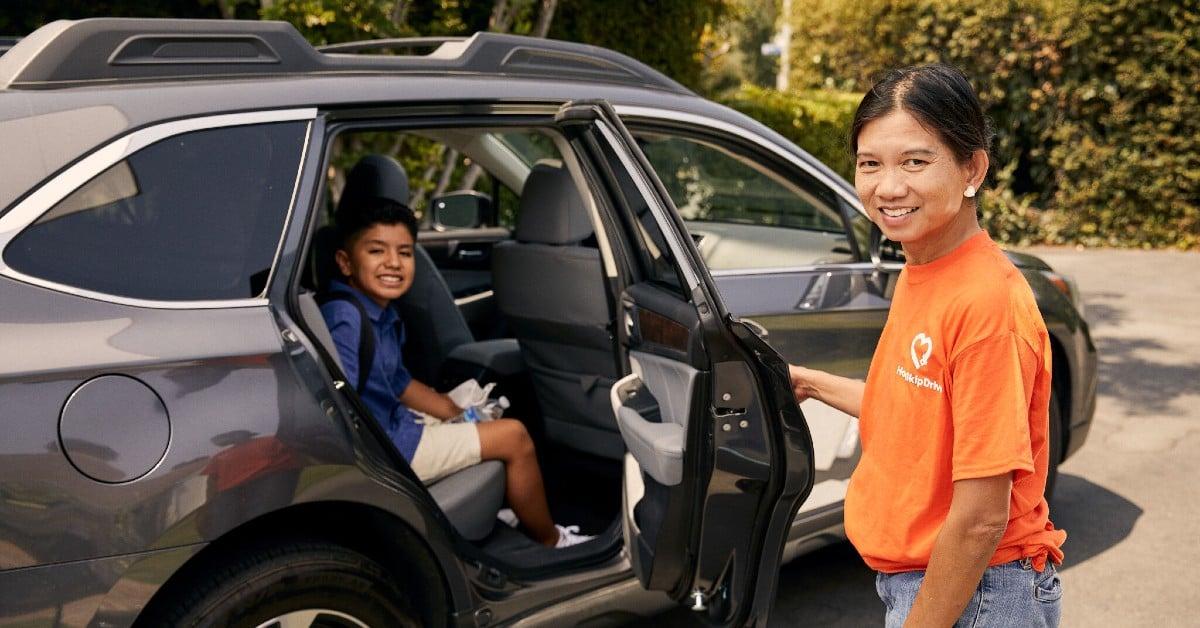 Driver in orange t-shirt opening car door for young student in school uniform sitting in back seat