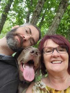 Cheerful selfie of two people and their happy dog with tongue out, taken outdoors among birch trees
