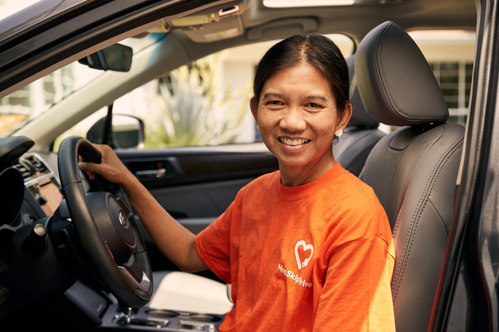 Service provider wearing orange Home Skip Drive t-shirt sitting in driver's seat of modern car, smiling warmly at camera