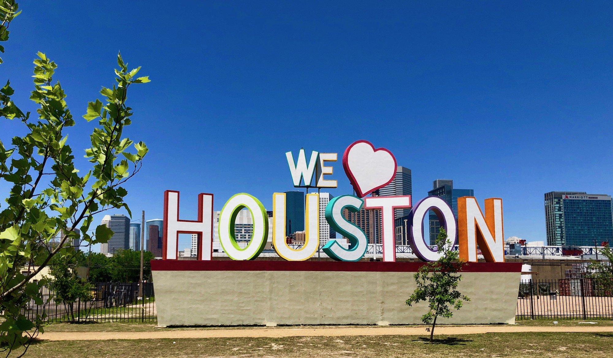 Colorful 'We Love Houston' sign with heart symbol against downtown Houston skyline on a clear blue day
