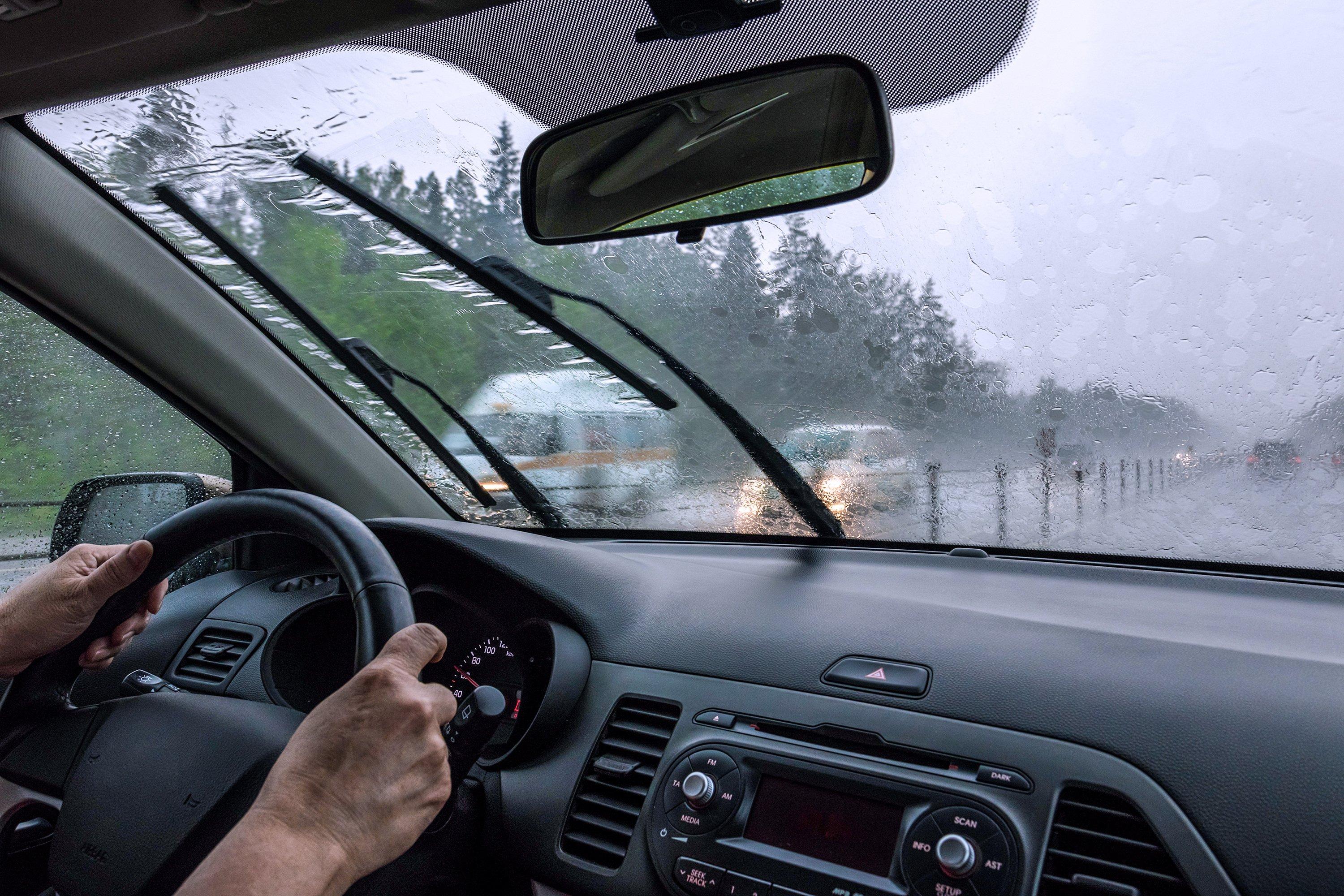 View through car windshield on rainy day with wipers on, showing dashboard and steering wheel with hands on wheel