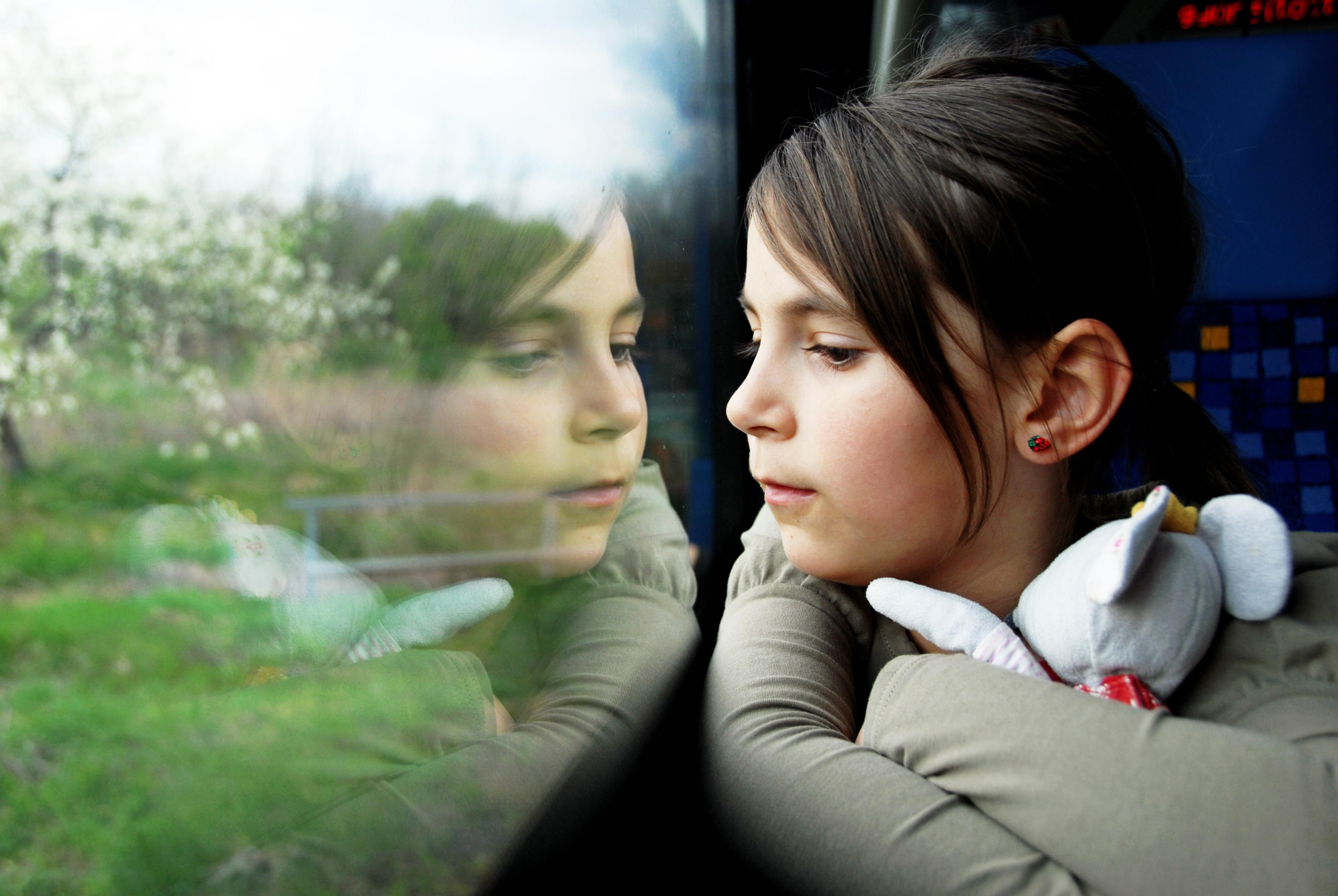 Young person gazing out train window with reflection, holding stuffed toy, creating contemplative double image effect