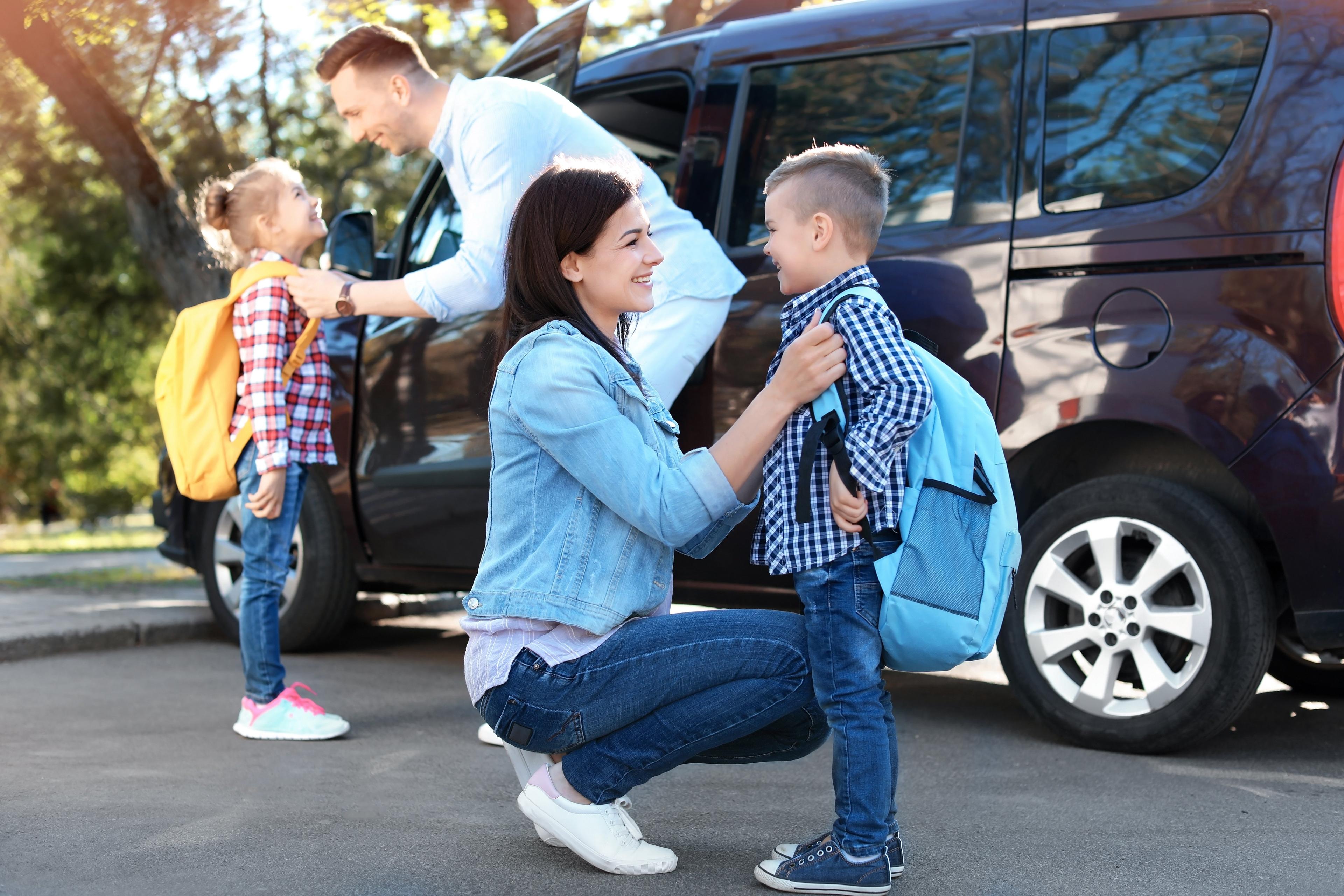 Parents helping children with backpacks near minivan during morning school drop-off, showing warm family interaction