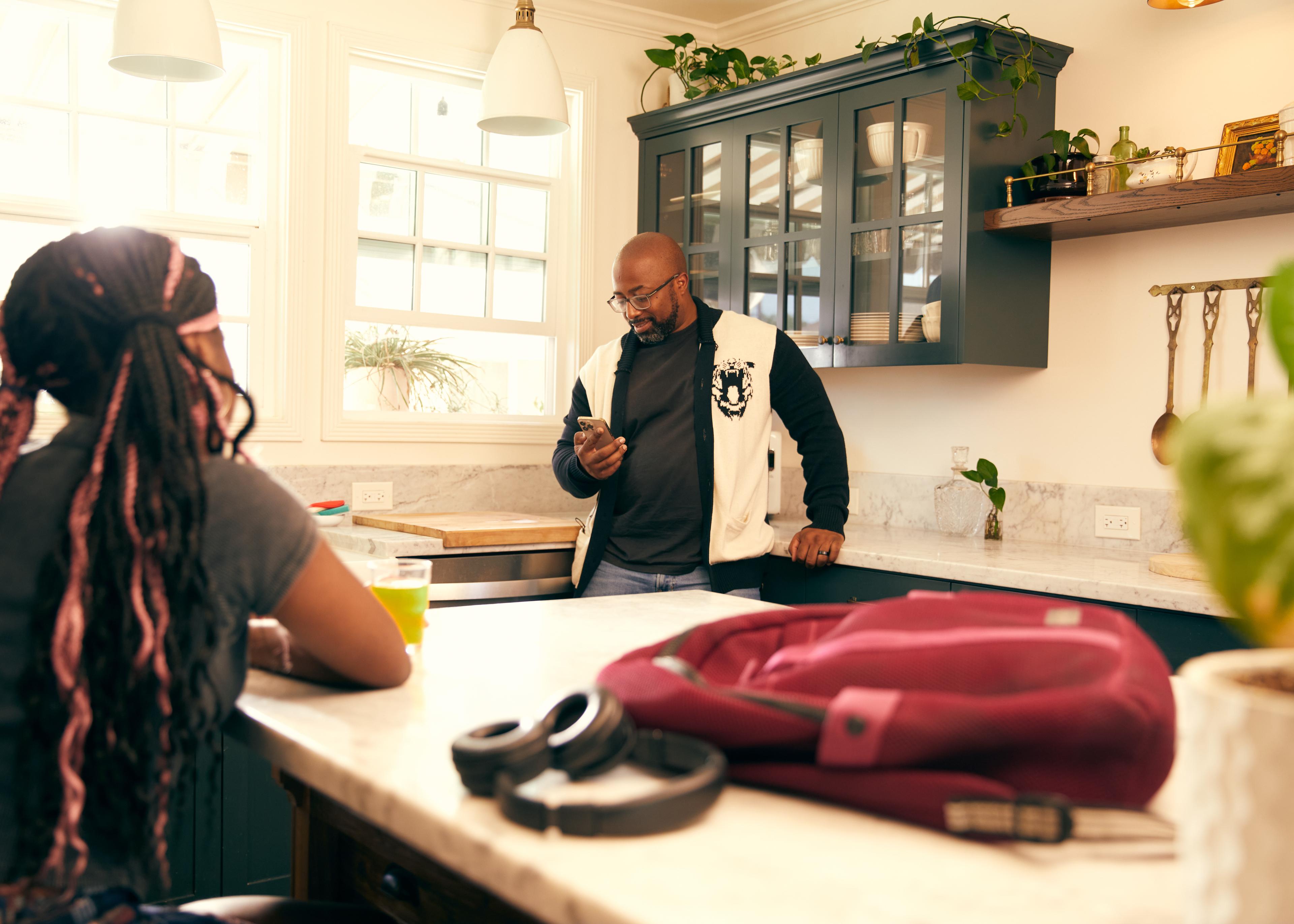 Two people chatting in a modern kitchen with headphones and backpack on counter, morning sunlight streaming through windows