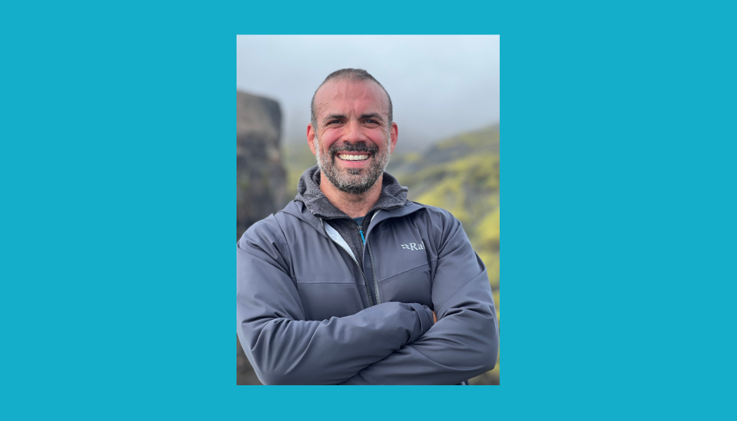 Portrait of a bearded man smiling warmly while wearing a grey Rab jacket outdoors with mountains in the background
