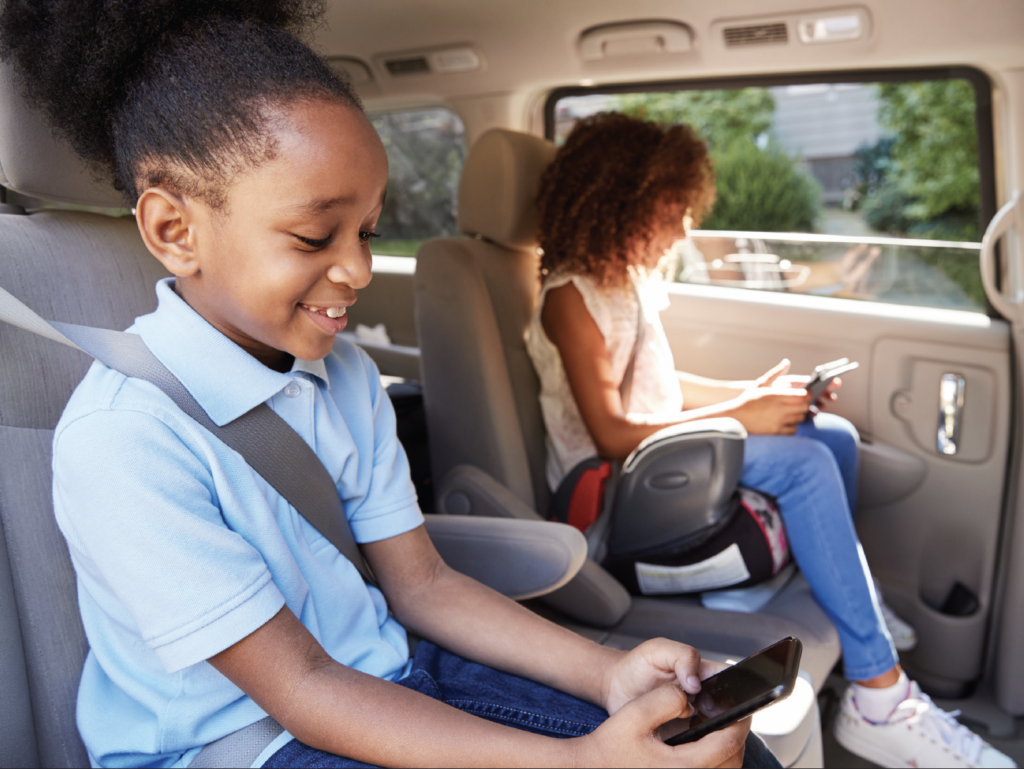 Two children sitting in car safety seats looking at mobile devices, wearing seatbelts and casual clothing