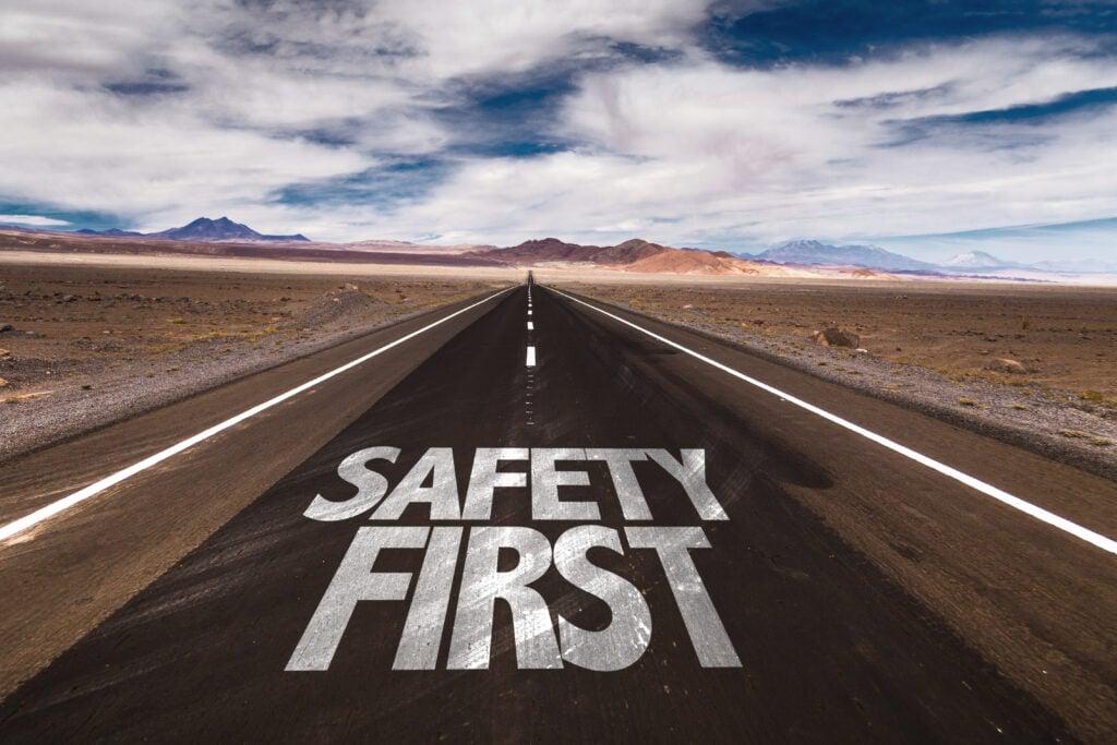 Text reading 'SAFETY FIRST' painted on an empty desert highway stretching toward distant mountains under cloudy blue sky