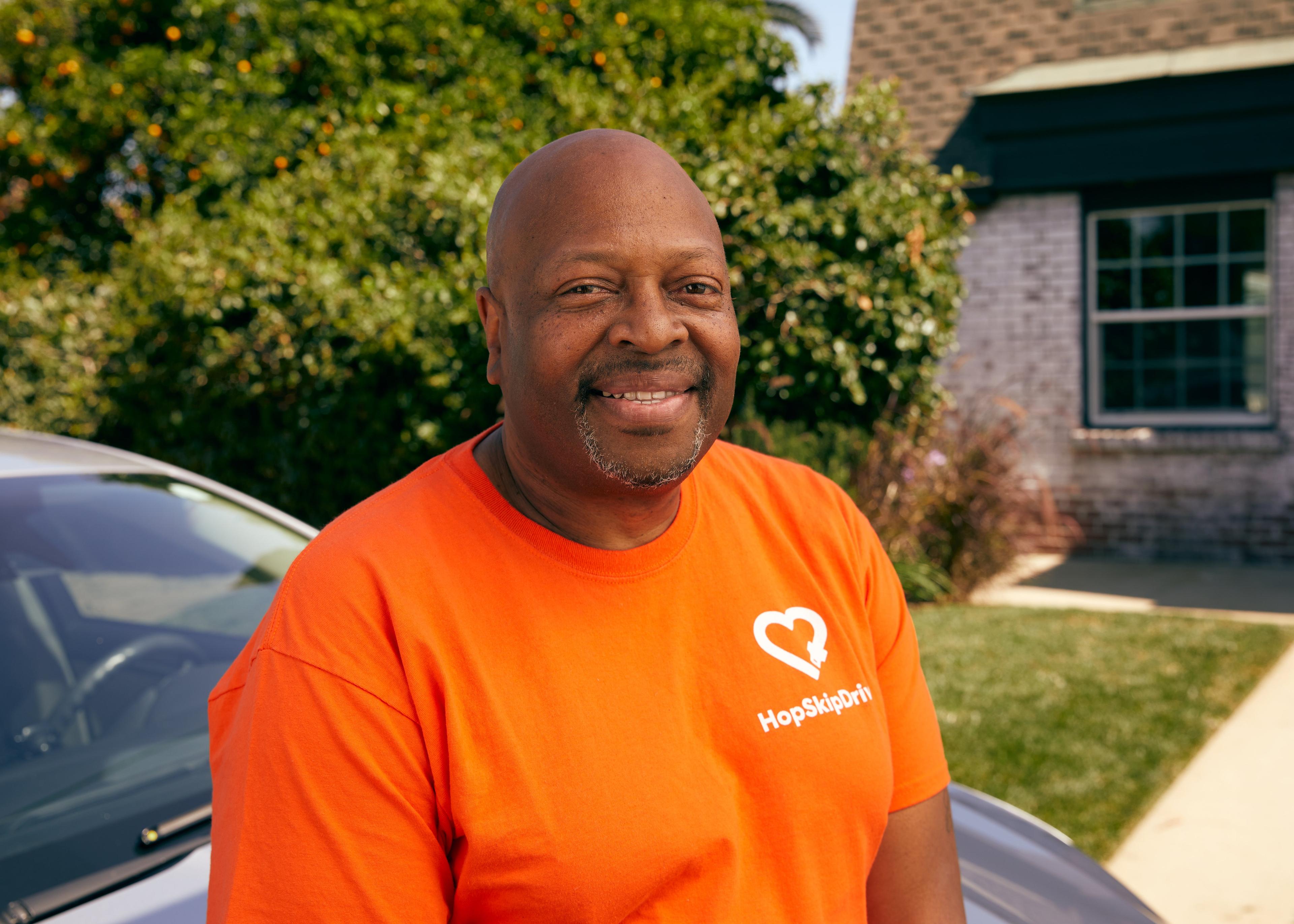Professional driver wearing bright orange HopSkipDrive t-shirt smiling warmly at camera in residential setting