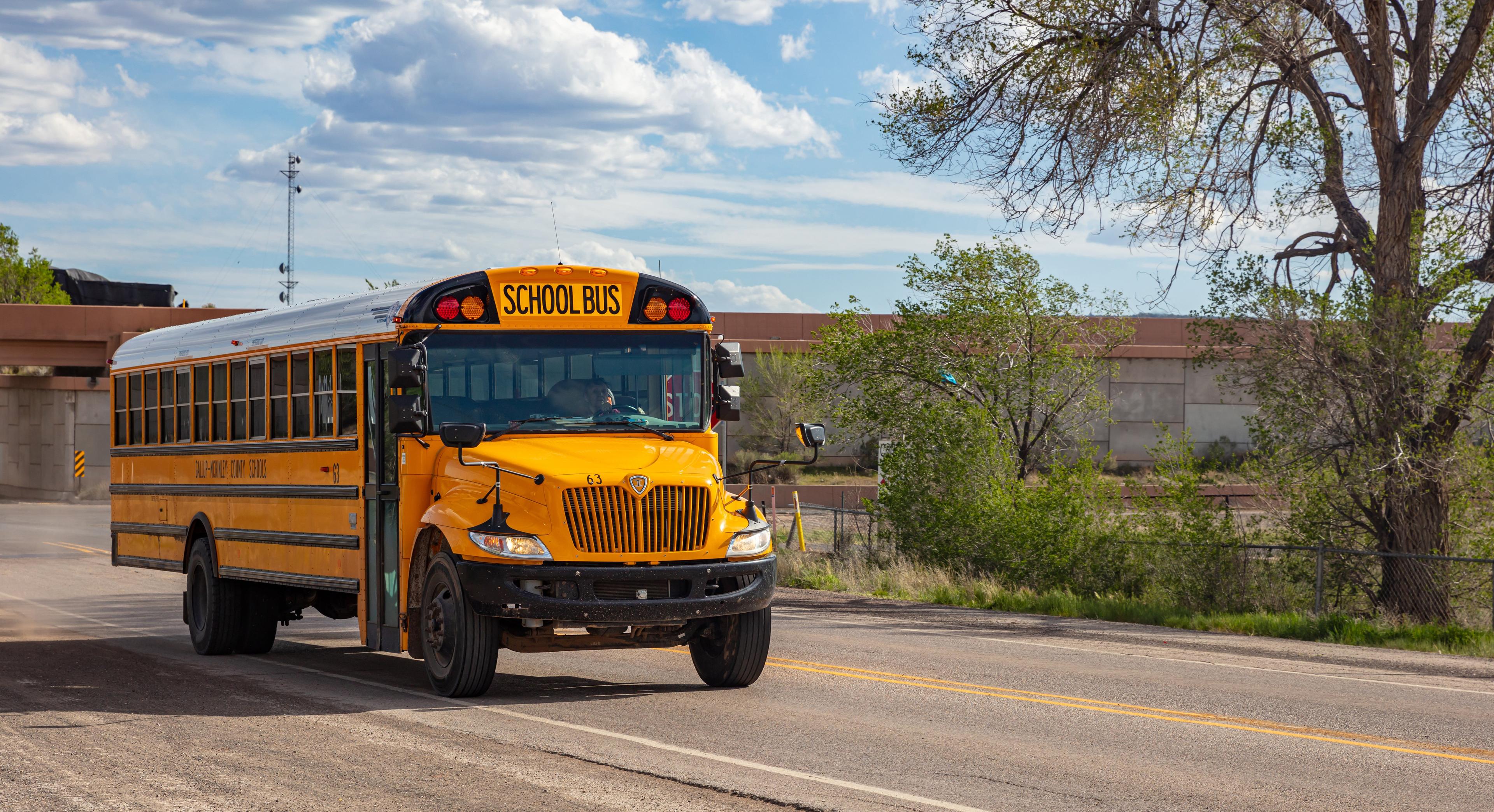 A bright yellow International school bus driving on a rural road with concrete barriers and scattered vegetation in the background
