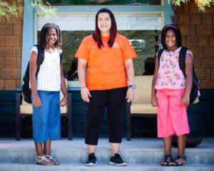 Three people in casual summer attire standing and smiling outside a building entrance with glass doors