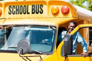 School bus driver standing in the doorway of a yellow school bus with a welcoming smile