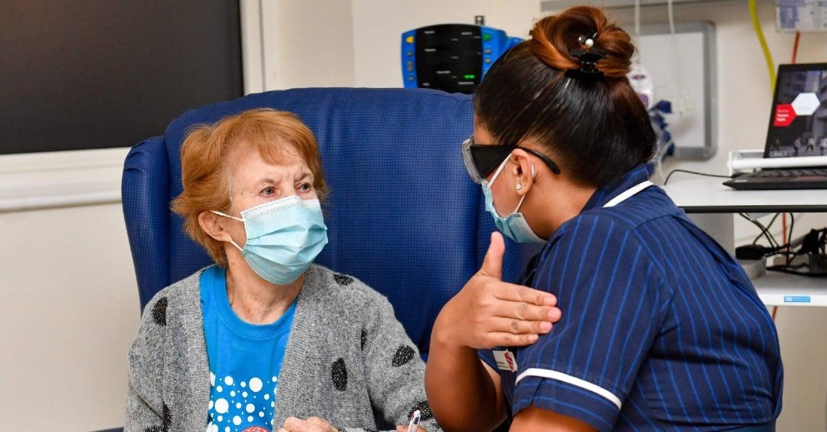 Healthcare worker in navy uniform talking with elderly patient in blue chair, both wearing protective face masks