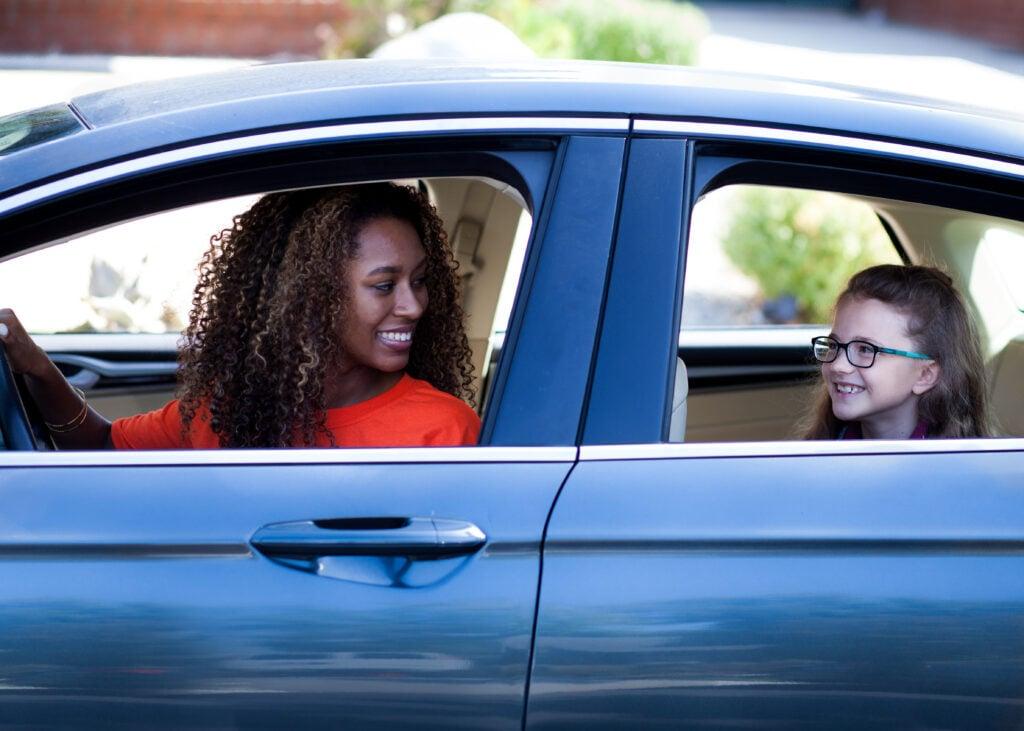 Two passengers in a blue car share a cheerful moment, driver in orange shirt and young passenger with glasses in back seat