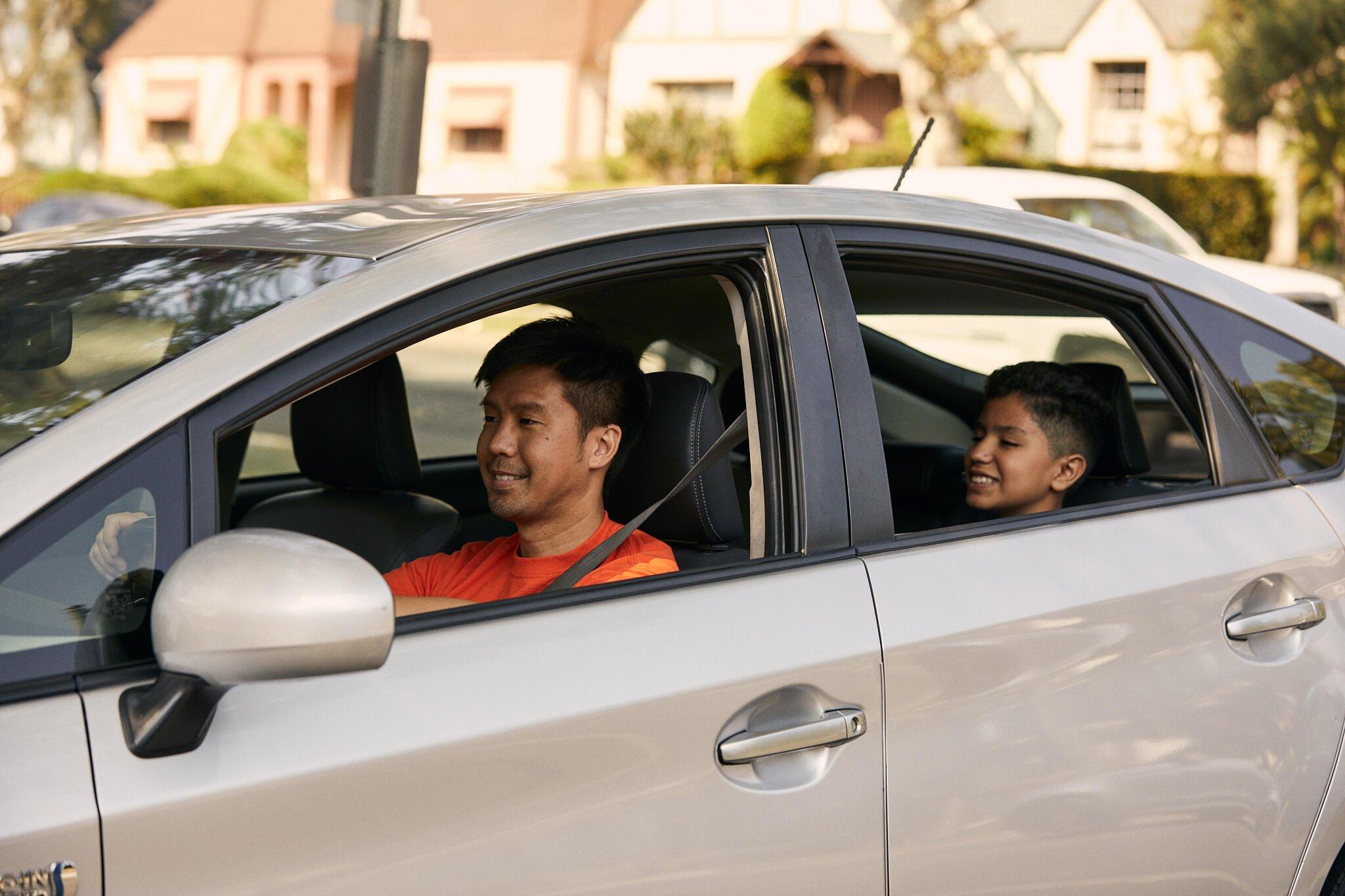 Parent in orange shirt driving car with young passenger in back seat, both smiling, in suburban setting