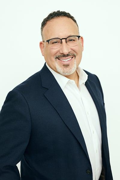 Professional headshot of a middle-aged man in navy blazer and white shirt, wearing glasses and smiling warmly at camera