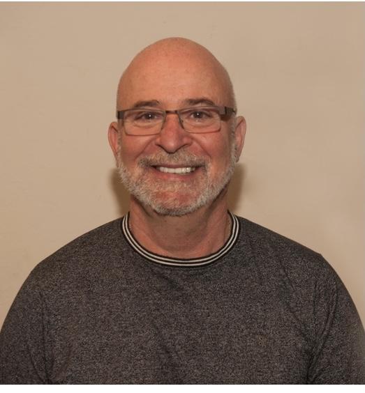 Friendly middle-aged man with glasses and gray beard wearing a gray sweater, smiling warmly at camera against neutral background