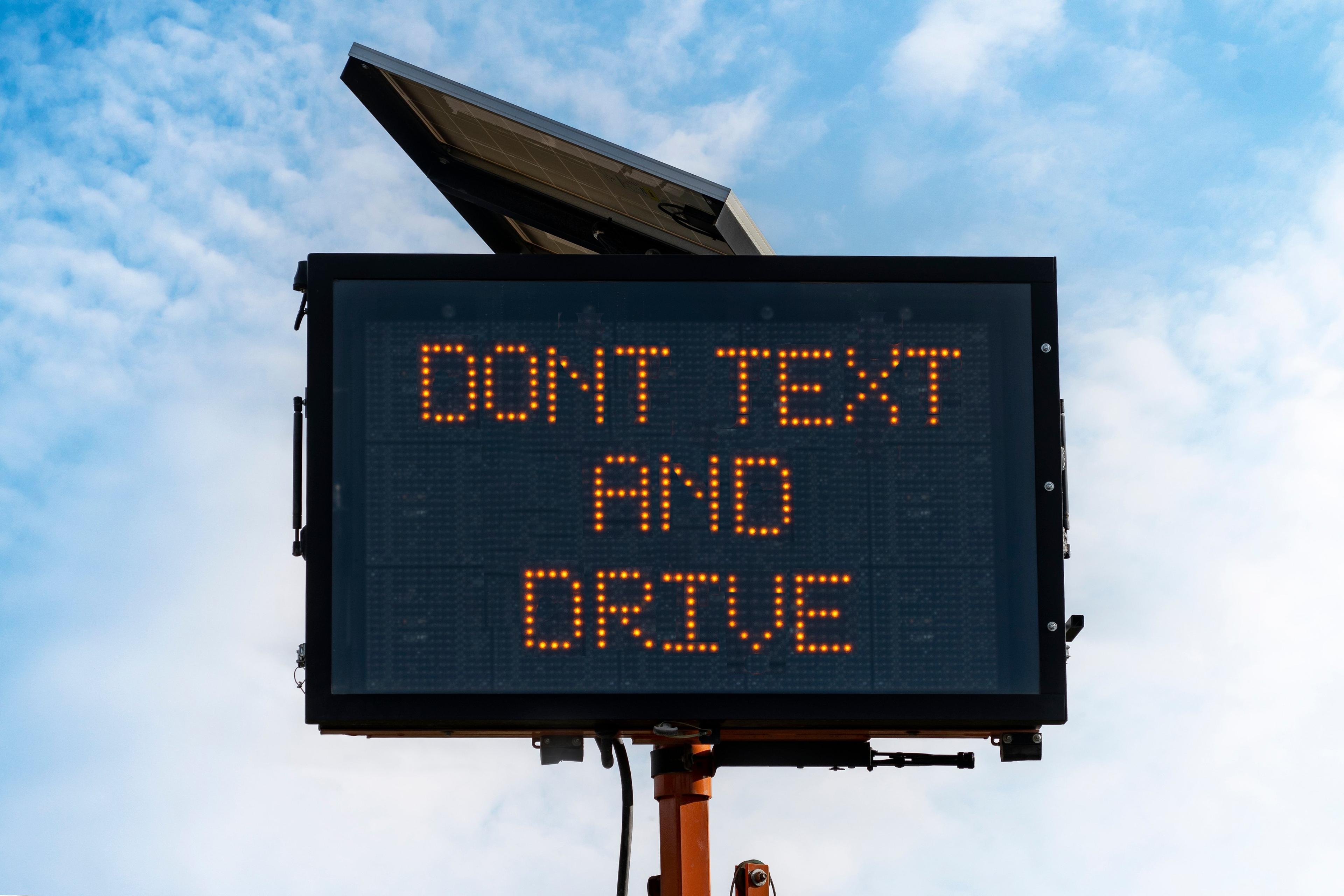 Digital traffic sign with orange LED text reading 'DONT TEXT AND DRIVE' against blue sky background