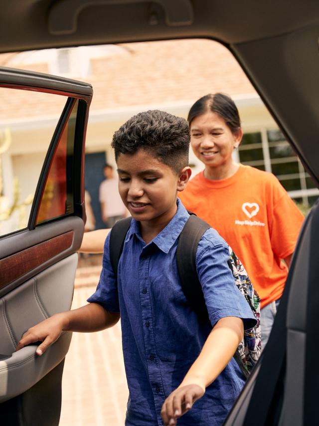 A student wearing a blue shirt and backpack enters a car while a volunteer in an orange t-shirt smiles in the background