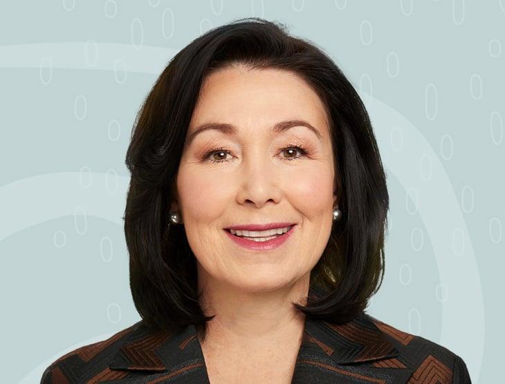 Professional headshot of an Asian woman in business attire smiling warmly, wearing pearl earrings against a light blue patterned background